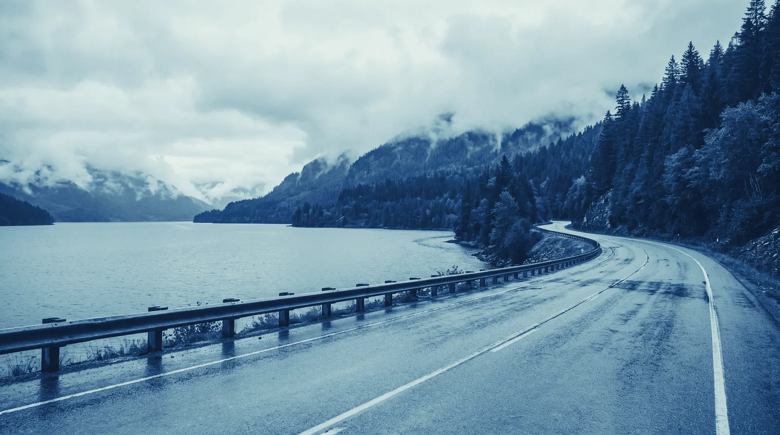 A computer-made picture of a two-lane highway curving along a grey mountain lake, with low cloud and forest on both shores